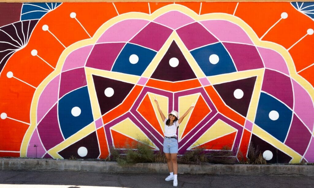 A smiling person stands in front of a mural of a colorful Rangoli. 